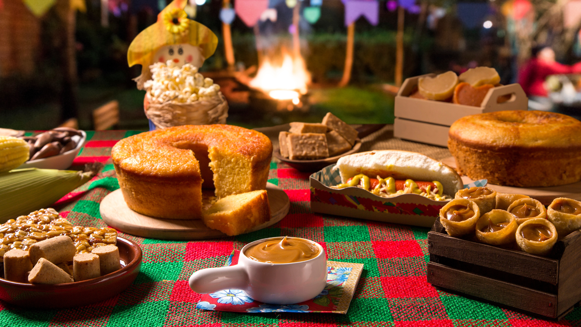 Mesa enfeitada com decorações de festa junina com comidas típicas da festa como bolo de milho, paçoca, cachorro quente, pipoca