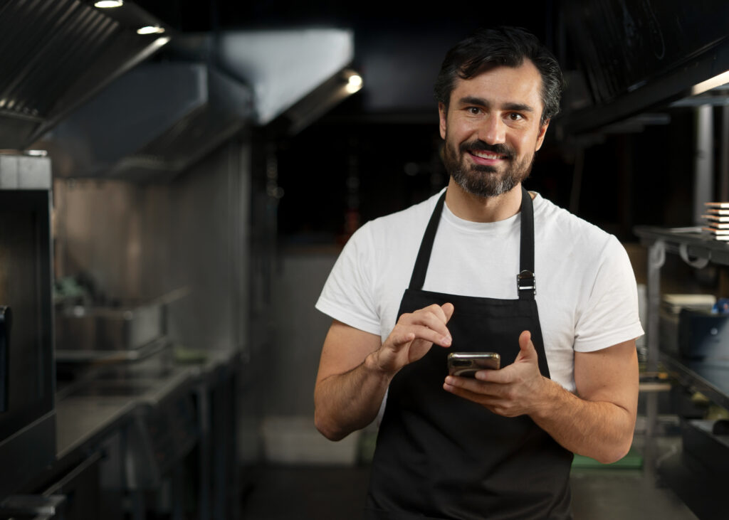 Um homem de cabelos escuros e pele clara está dentro de uma cozinha de restaurante segundando um celular nas mãos