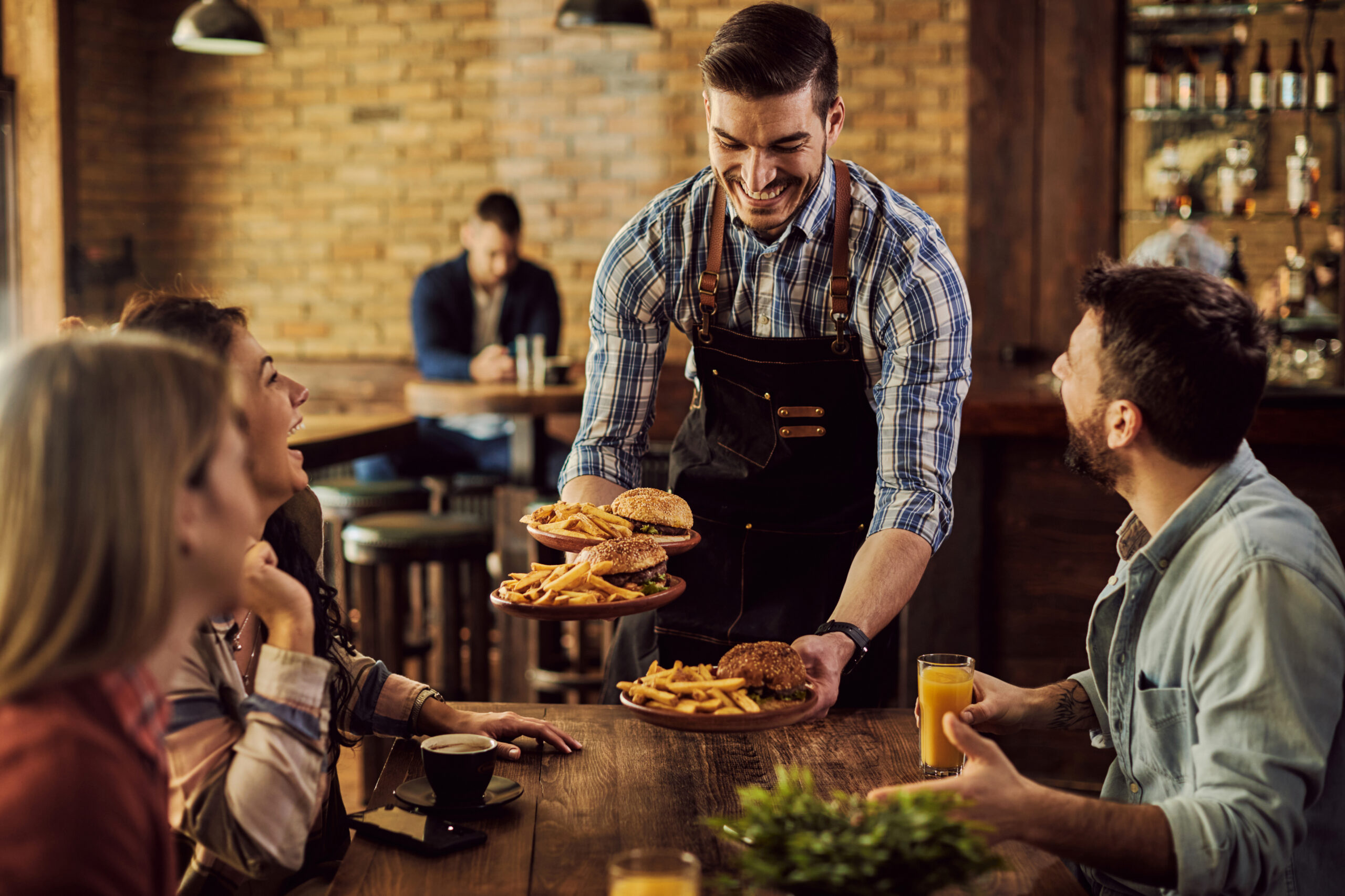 Um grupo de amigos está se divertindo enquanto um garçom está servindo pratos com hamburguer e batata frita em um restaurante. O foco da imagem está no garçom.