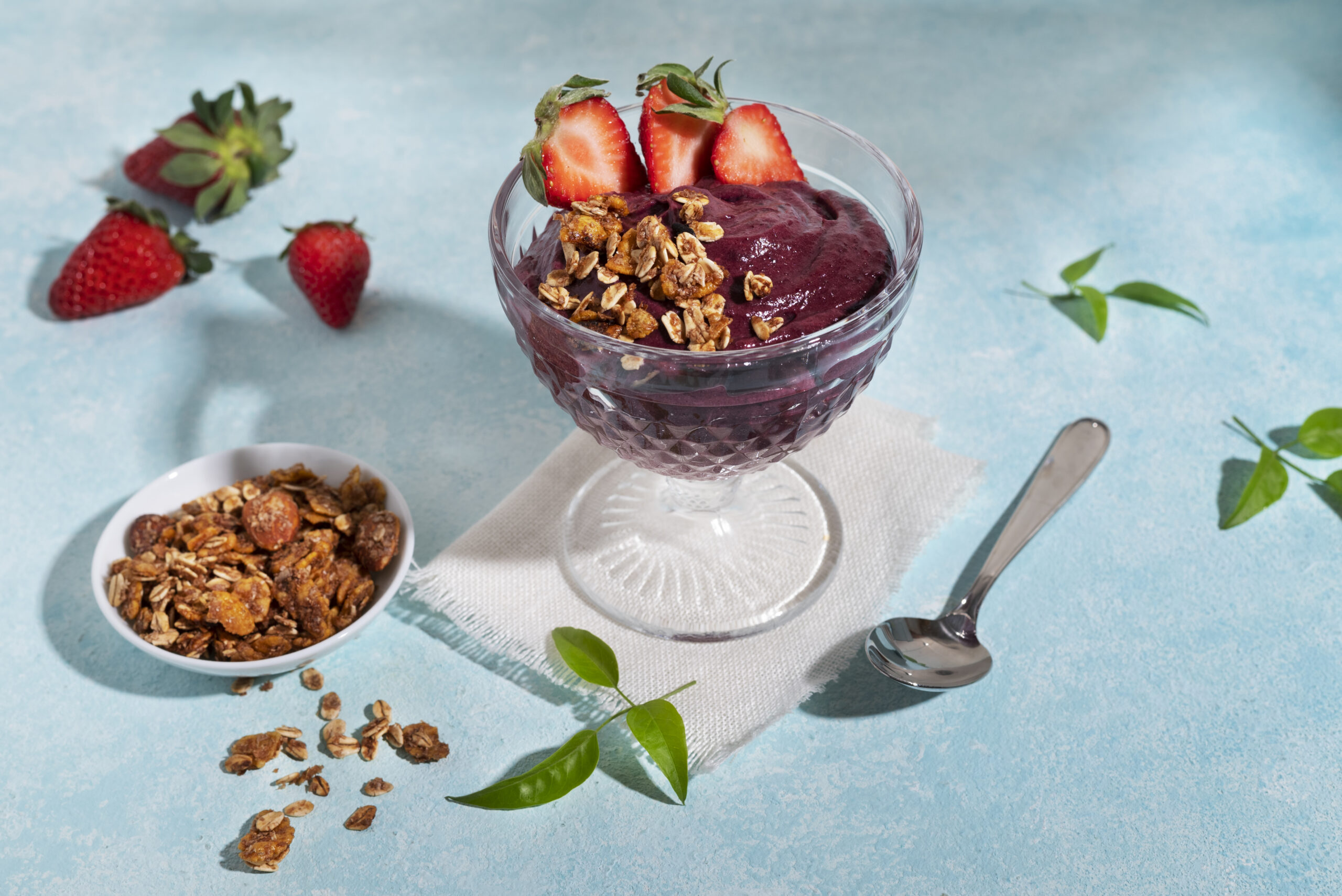 Taça de açaí com morango e granola em cima de uma toalha azul clara com colher ao lado