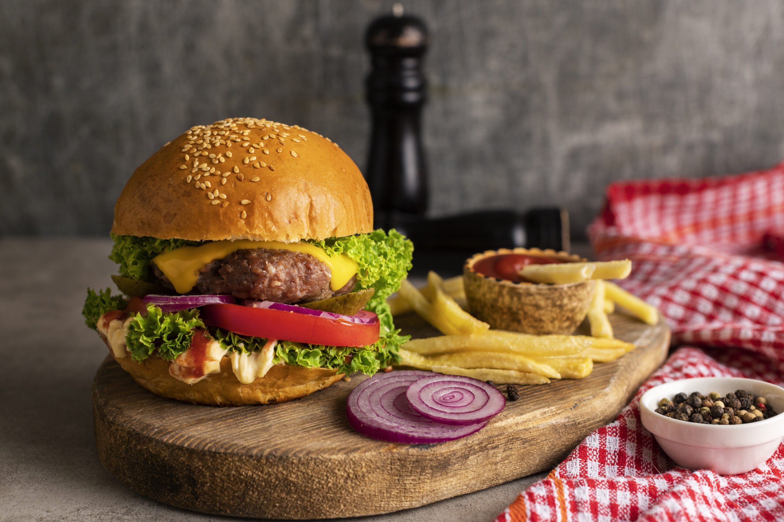 Hamburguer com queijo, tomate, cebola e alface em cima de uma tábua com batata frita e outros ingredientes ao lado