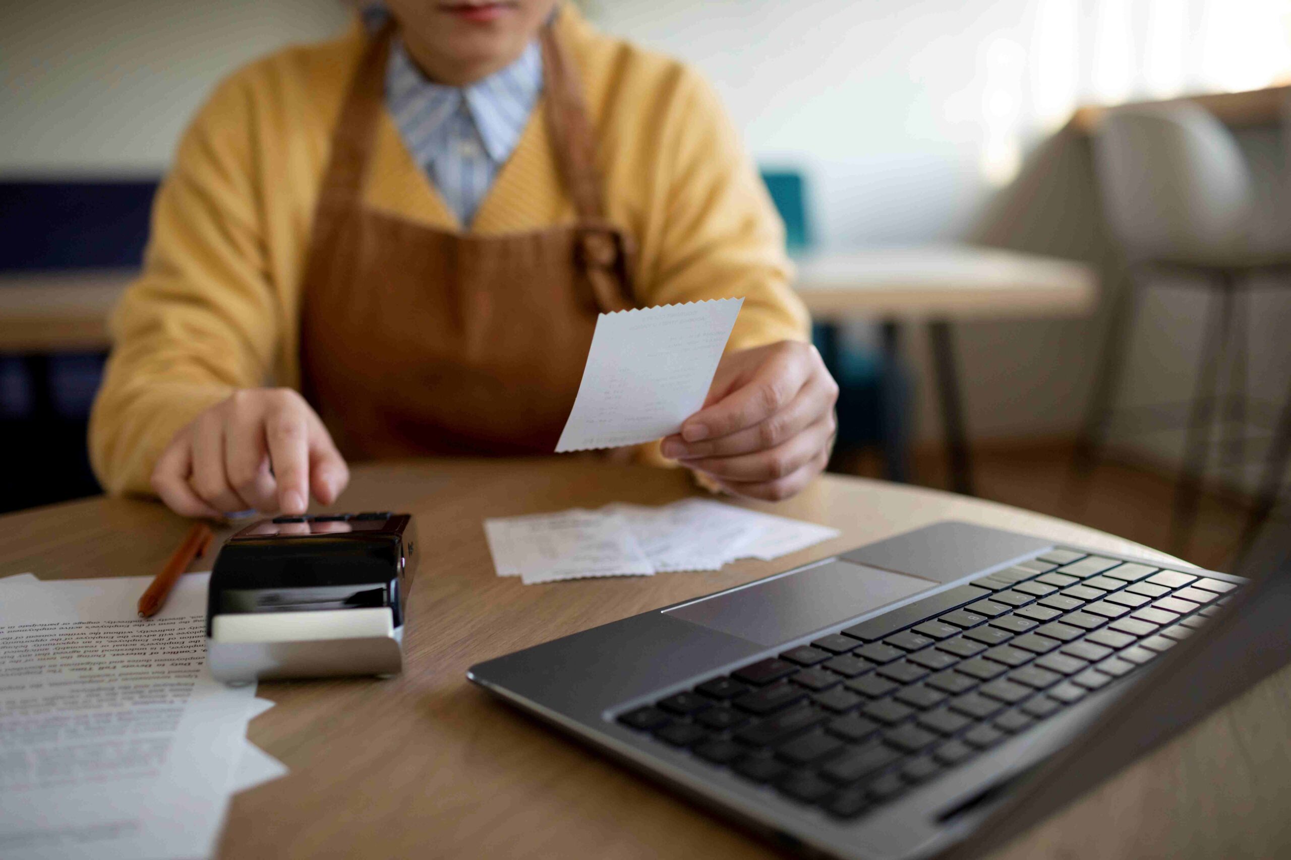 Pessoa fazendo contas mexendo em uma maquininha de cartão enquanto analisa cupom fiscal